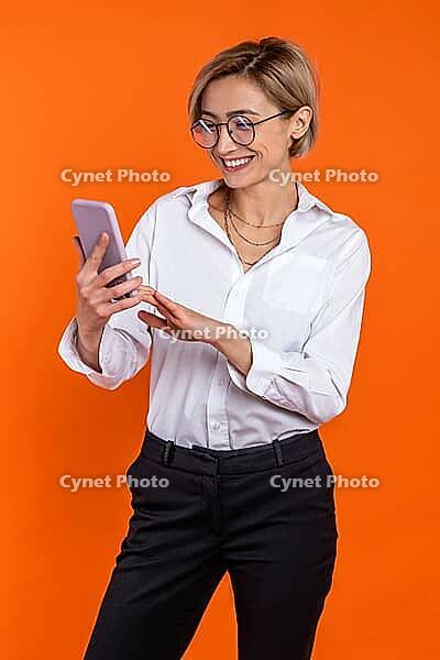 Joyful beautiful woman wearing white official style shirt using mobile phone isolated over orange background [IBR123569293]