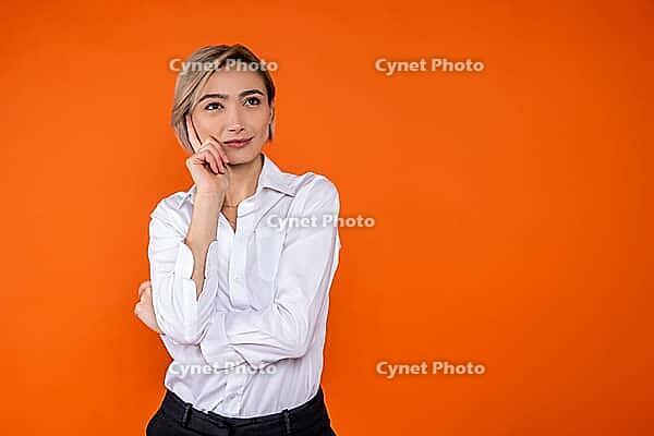 Pensive woman wearing white official style shirt thinking holding chin free space isolated over orange background [IBR123569296]