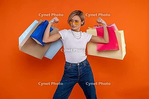 Funny strong woman in white t shirt and jeans raised arms with shopping bags isolated over orange background [IBR123569299]