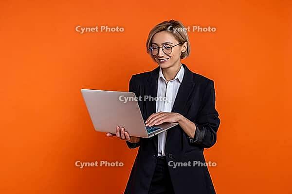 Confident businesswoman wearing black official style suit working on laptop in office isolated over orange background [IBR123569301]