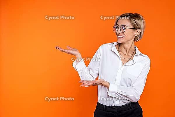 Delighted woman wearing white official style shirt presenting copy space on palm isolated over orange background [IBR123569302]