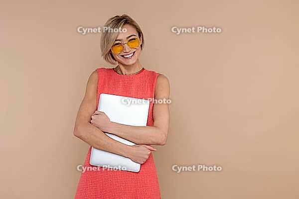 Confident happy beautiful woman in dress and sunglasses holding laptop empty space isolated over beige background [IBR123569303]
