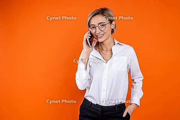 Smiling woman wearing white official style shirt talking on smartphone isolated over orange background [IBR123569306]