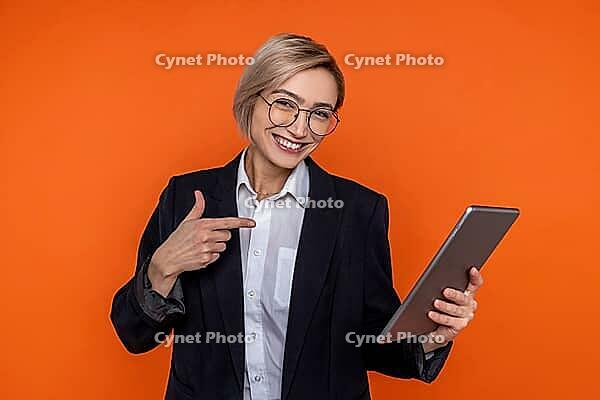 Happy woman wearing black official style suit pointing at tablet isolated over orange background [IBR123569307]
