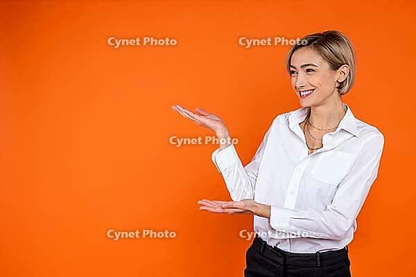 Satisfied woman wearing white official style shirt showing empty space on palm isolated over orange background [IBR123569308]
