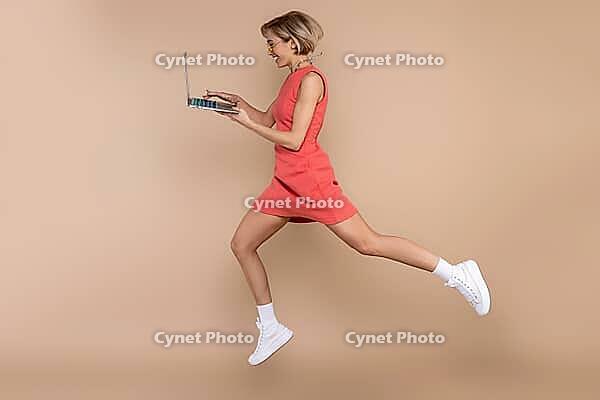 Jumping woman in dress and sunglasses using laptop working online isolated over beige background [IBR123569311]