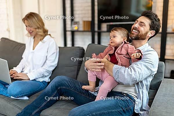 Father carrying sleeping daughter while looking at woman using laptop on sofa [IBR123569319]