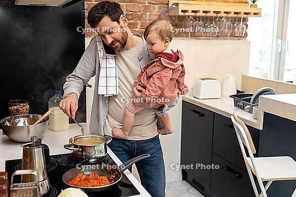 Busy father looking after baby girl making meal and talking on mobile phone, multitasking man at home in kitchenn [IBR123569320]