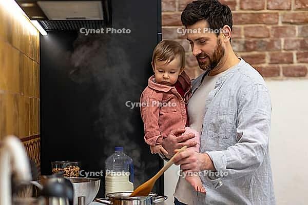 Father cooking meal holding toddler daughter in kitchenn preparing soup [IBR123569322]