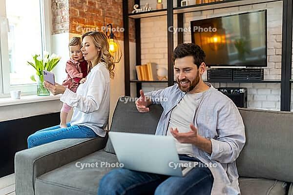 Father working on laptop, mother and daughter on background making selfie on phone in home interior [IBR123569323]