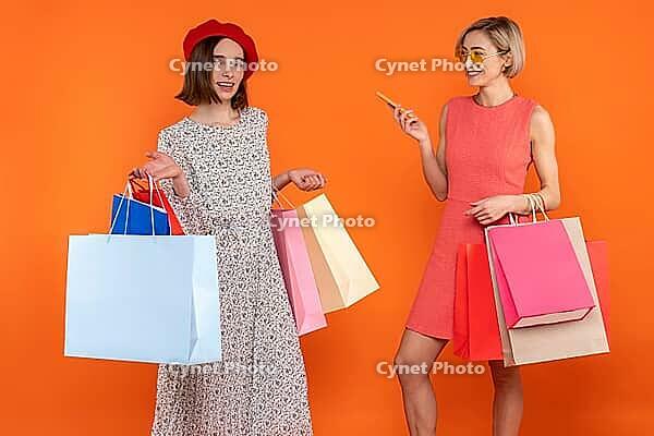 Satisfied women friends with shopping bags enjoying new purchases isolated over orange background [IBR123569324]