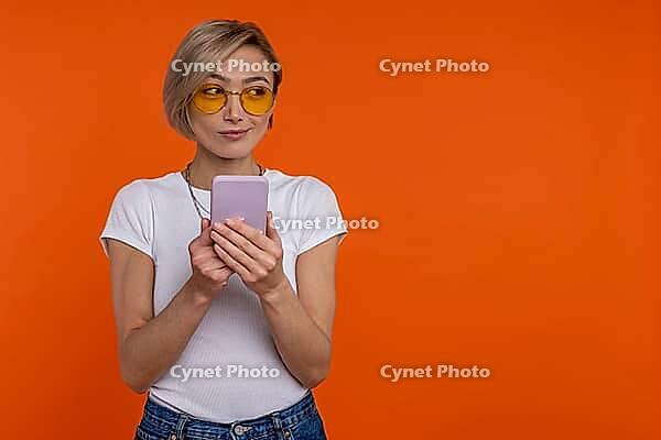 Cunning sly woman wearing white T-shirt using mobile phone checking social networks isolated over orange background [IBR123569325]