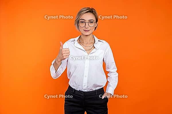 Attractive confident woman wearing white official style shirt showing thumb up isolated over orange background [IBR123569326]