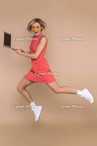 Jumping woman in dress and sunglasses using laptop working online isolated over beige background [IBR123569327]