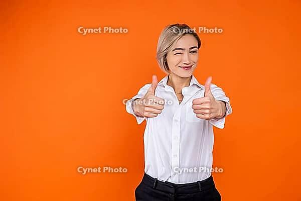 Positive woman wearing white official style shirt showing like gesture and winking to camera isolated over orange background [IBR123569328]