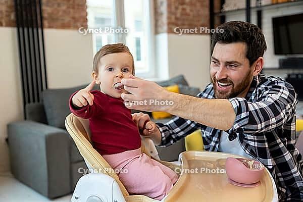 Bearded father feeding little baby daughter sitting in highchair with puree or porridge by spoon at home [IBR123569331]