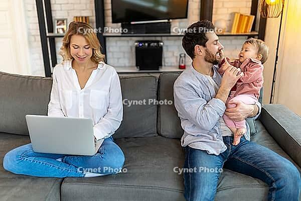 Father carrying infant daughter while mother using laptop on sofa remote work husband helping his wife with baby child [IBR123569334]