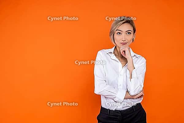 Pensive thoughtful woman wearing white official style shirt thinking holding chin isolated over orange background [IBR123569336]