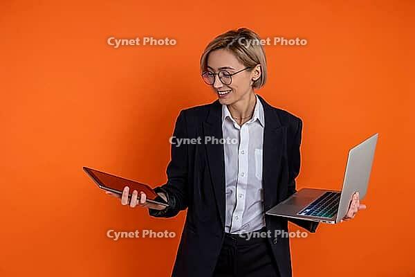 Joyful woman wearing black official style suit choosing laptop or tablet isolated over orange background [IBR123569337]