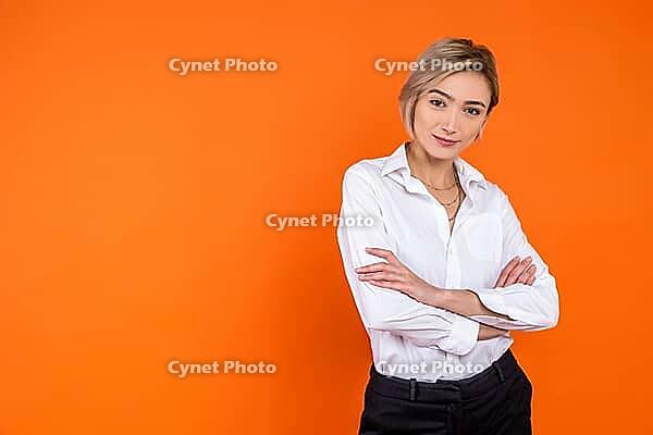 Confident woman wearing white official style shirt standing with crossed arms looking at camera isolated over orange background [IBR123569339]
