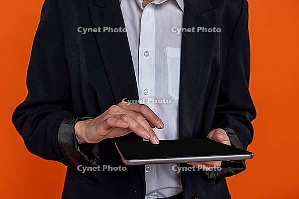 Unrecognizable woman wearing black official style suit using tablet isolated over orange background [IBR123569341]