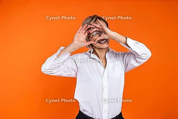 Romantic woman wearing white official style shirt looking through heart shape with hands isolated over orange background [IBR123569342]