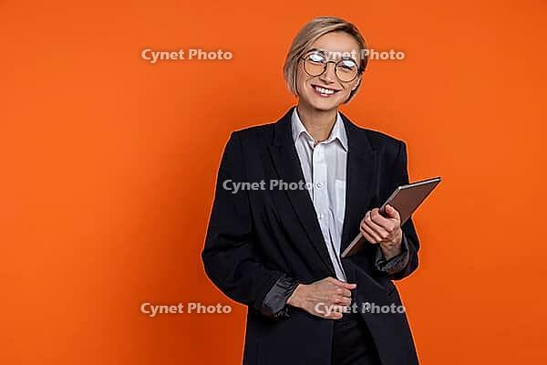 Confident satisfied woman wearing black official style suit standing with tablet looking at camera isolated over orange background [IBR123569343]