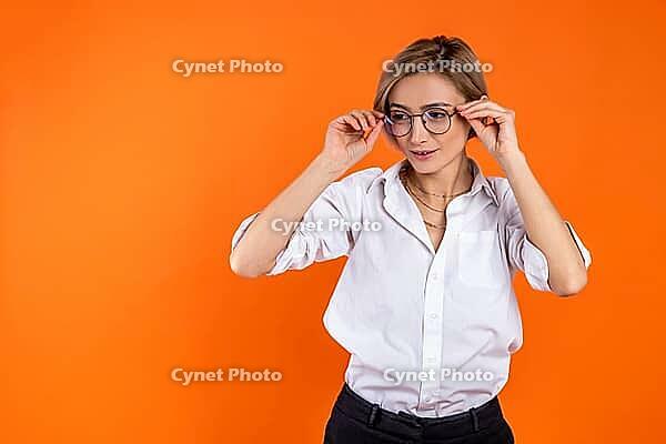 Attractive woman wearing white official style shirt holding frame of glasses looking away isolated over orange background [IBR123569344]