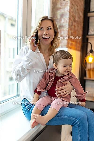 Smiling cheerful woman sitting on windowsill in living room using mobile phone holding baby and talking on phone [IBR123569345]