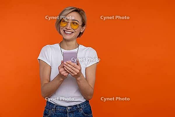 Positive woman wearing white T-shirt using mobile phone checking social networks isolated over orange background [IBR123569346]