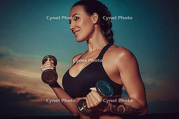 Young sports woman training with dumbbells on twilight sky background, tattoo on hand [IBR123569355]