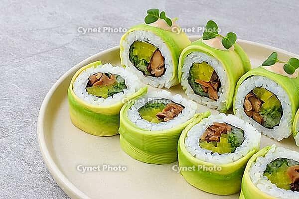 Food, A sushi roll wrapped in thin avocado slices, filled with marinated mushrooms, crisp pickled radish, and seasoned rice, topped with spicy mayo and microgreens, served on a neutral ceramic plate [IBR123622504]