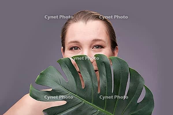 Smiling caucasian adult female peeking from monstera leaf against gray background [IBR123629728]