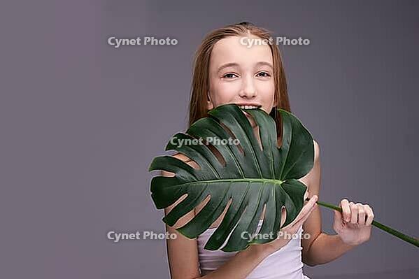 Young caucasian female teen with long hair holding large monstera leaf against gray background and smiling [IBR123629729]