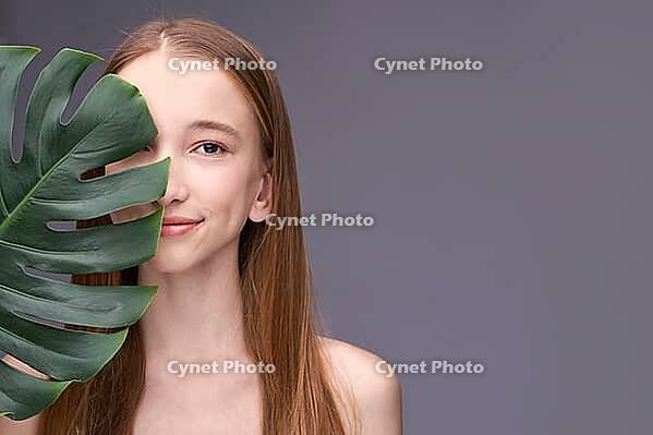 Young caucasian female with long hair holding leaf in minimalist portrait on gray background [IBR123629730]