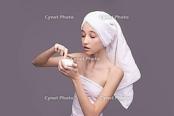 Young asian female applying skincare cream with towel on head against gray background [IBR123629732]