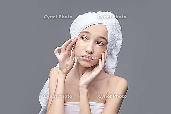 Young asian female applying under eye patches for skincare routine with towel wrapped around head [IBR123629735]