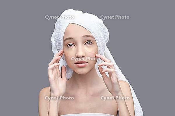 Young caucasian female with under eye patches and towel on head, gray background, skincare routine [IBR123629736]