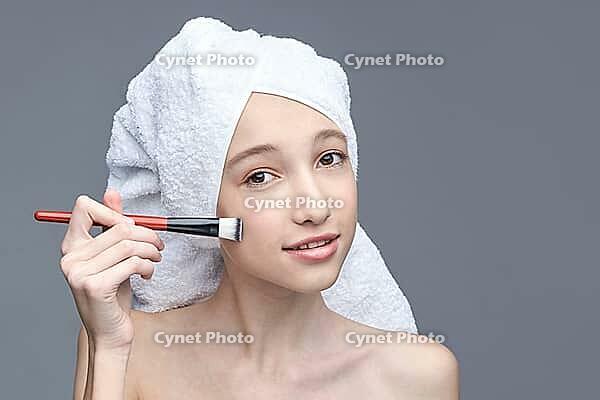 Young caucasian female applying makeup with brush and towel wrap against gray background [IBR123629739]