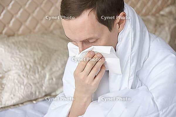 Caucasian adult male with cold or allergies blowing nose into tissue in bedroom wearing white robe [IBR123629740]