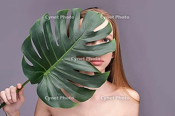 Young caucasian female partially covered by large green leaf against gray background [IBR123629745]