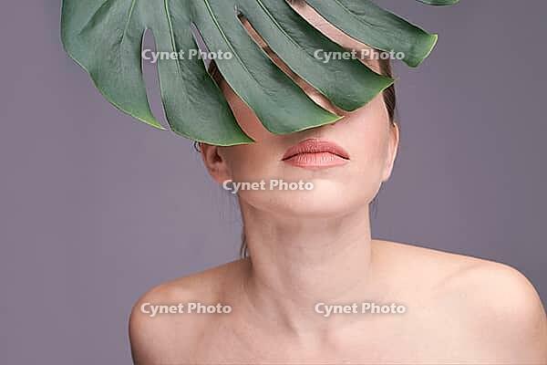 Young caucasian female partially covered by monstera leaf with neutral expression in studio setting [IBR123629748]