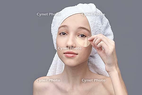 Young caucasian female applying eye patch with towel wrapped hair in skincare routine on grey background [IBR123629749]