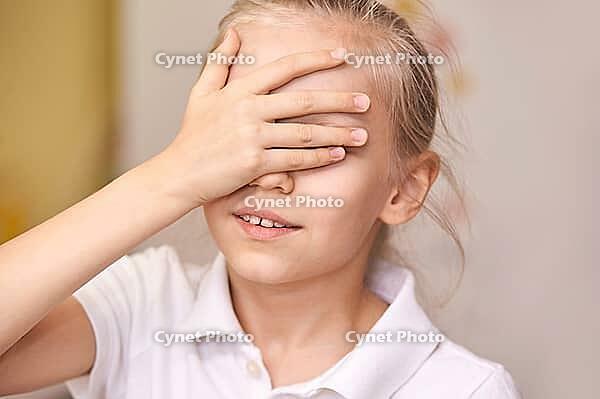 Young caucasian girl covering eyes with hand wearing white polo shirt indoors [IBR123629757]