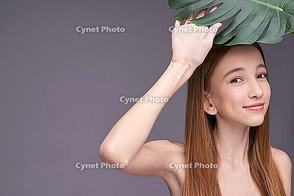Young caucasian female holding leaf with playful smile in minimalist setting [IBR123629758]