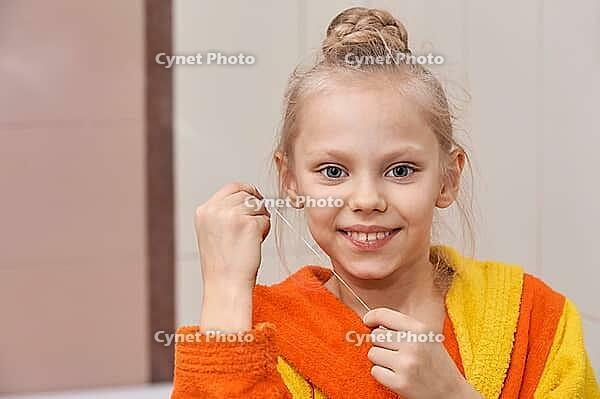 Smiling caucasian female child in bright bathrobe practicing dental hygiene with floss in bathroom setting [IBR123629759]