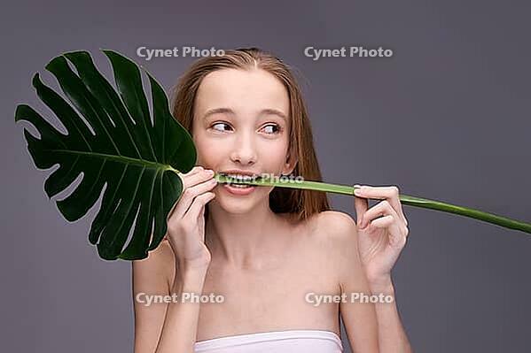 Young caucasian female playfully biting monstera leaf against grey background [IBR123629760]