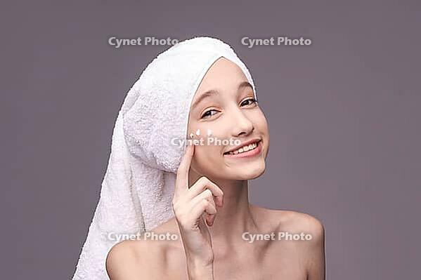 Young caucasian female applying skincare cream with towel wrap on head smiling [IBR123629761]