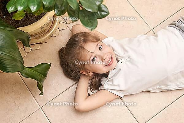 Smiling caucasian young girl relaxing on tiled floor next to potted green plants indoors [IBR123629767]