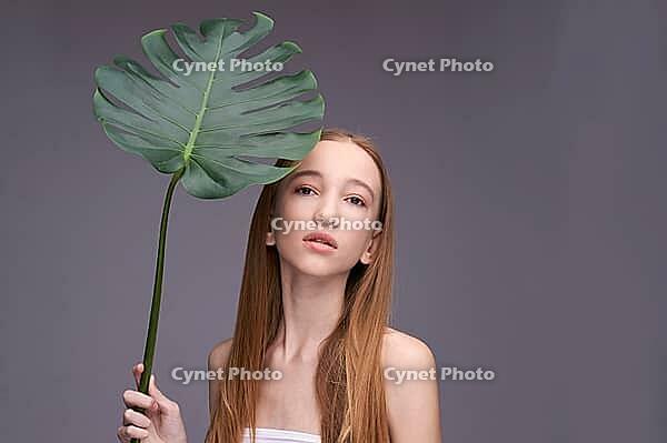 Young caucasian teen female holding large leaf against gray background with long straight hair and natural expression [IBR123629770]